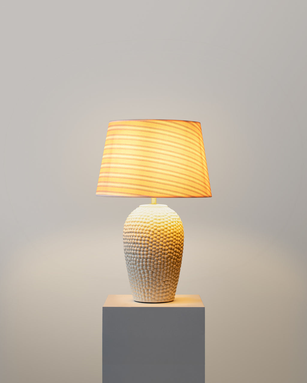 Table lamp with textured base and striped shade on a gray pedestal against a gray background