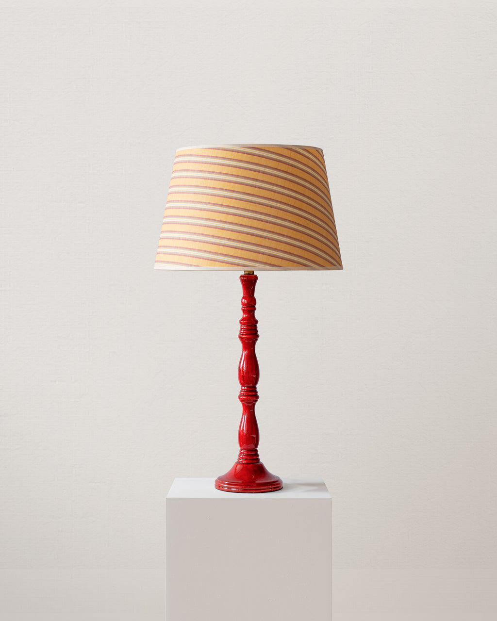 Table lamp with a red base and striped yellow shade on a white pedestal against a light gray background