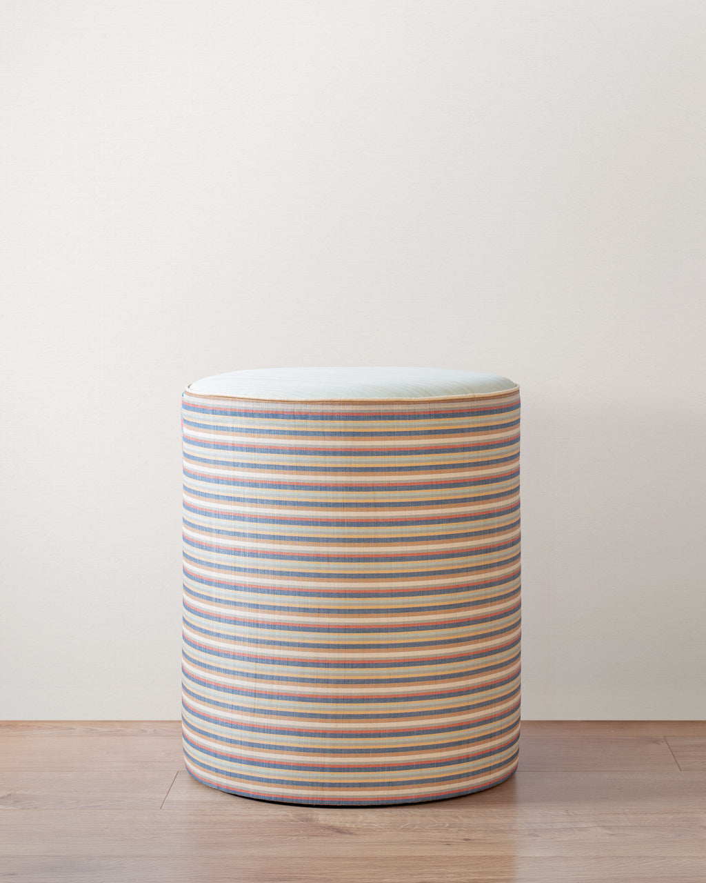 Cylindrical stool with multicolored stripes on a wooden floor against a plain wall.